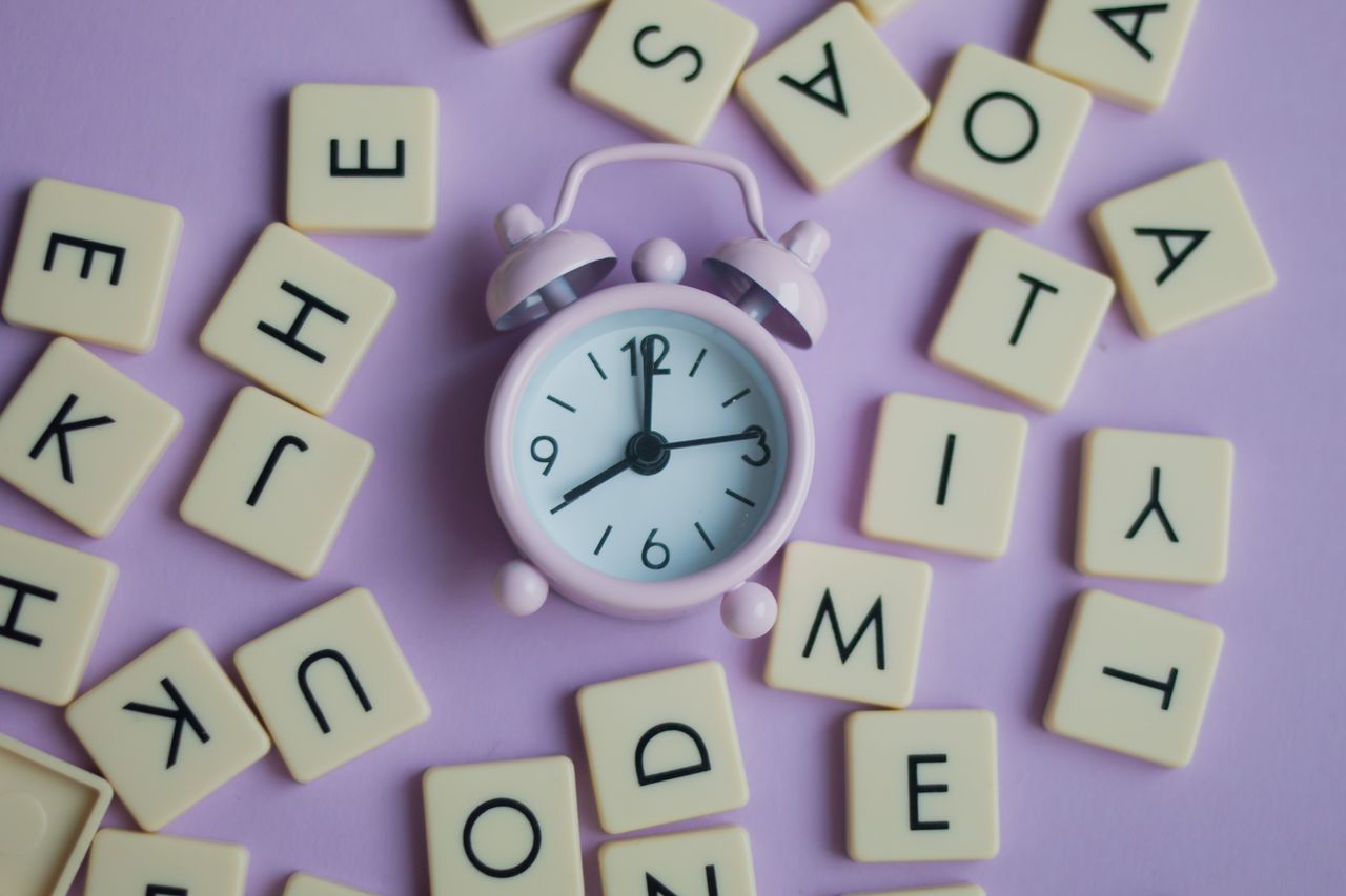 A minimalist photo of a pink analog alarm clock surrounded by letter tiles on a pastel pink surface.
